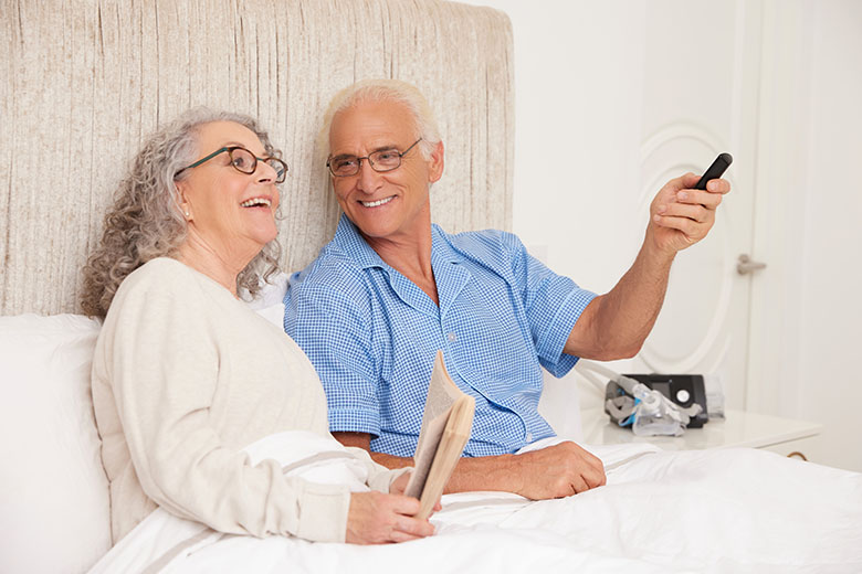 SleepApneaWhiteOlderCoupleSmilingSittingUpInBedHoldingRemoteReadingLifestyleResMedAirSense10