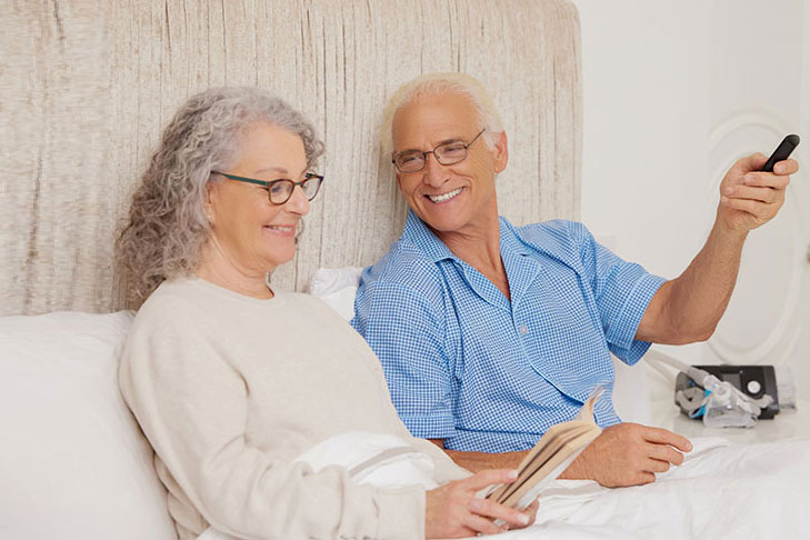 Sleep Apnea White Older Couple Smiling Sitting Up in Bed Holding Remote Reading Lifestyle ResMed AirSense10PAPMaskc