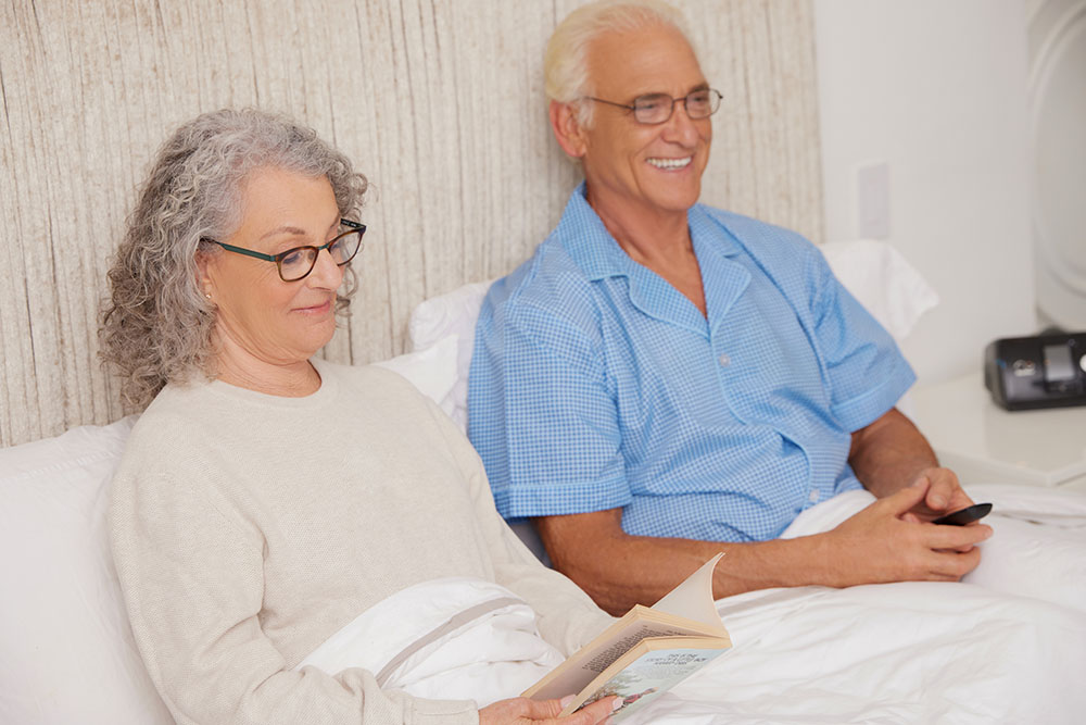 Sleep Apnea White Older Couple Smiling Sitting Up in Bed Holding Remote Reading Lifestyle