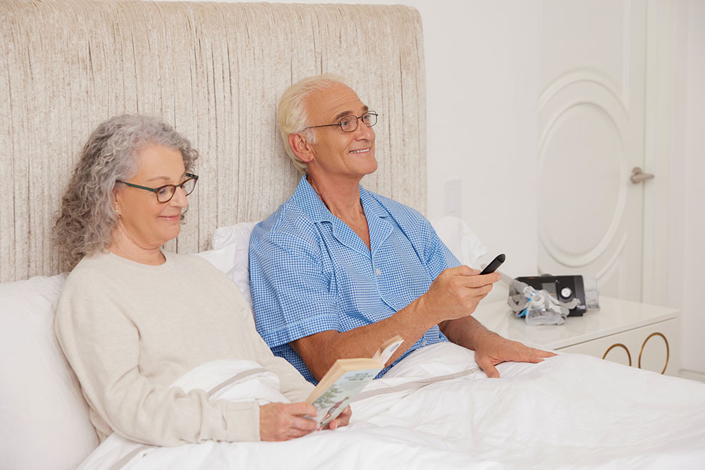 Sleep Apnea White Older Couple Smiling Sitting Up in Bed Holding Remote Reading Lifestyle 